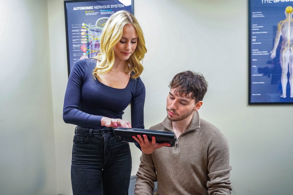 A chiropractic clinic staff member discusses patient health information on a tablet with a seated male patient in a consultation room.